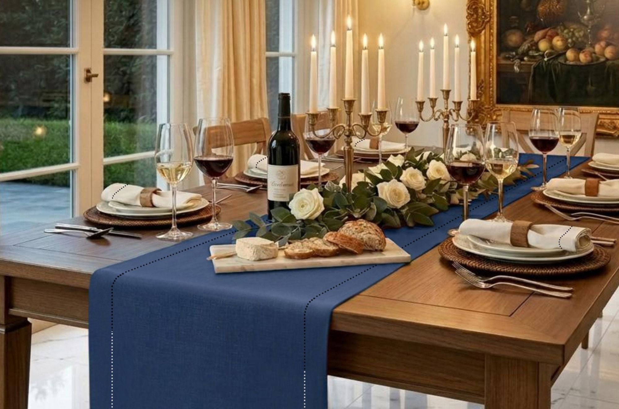 A styled dining table set for six with a navy blue linen table runner, dinner napkins, white roses, candles, wine glasses, bread, cheese, and a dark wine bottle in a warm, elegant dining room.